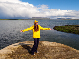 Person in bright yellow raincoat stands alone on a stone pier, arms wide open, facing a dramatic sky. Dark storm clouds swirl above calm water. Sligo, Ireland, Travel and tourism. © mark_gusev