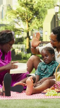 Vertical video: Parents and curious boy read a book together on a picnic blanket, sharing a learning and storytelling moment beside food and a picnic basket in an urban park during weekend family fun.