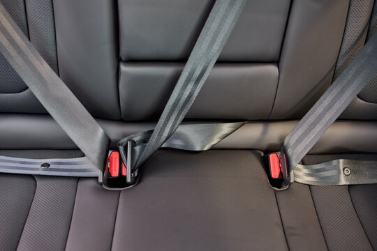 Close view of seatbelt buckle latched into car seat receiver on black leather upholstery with textured webbing, stitched trim, red accent detail, and soft directional lighting