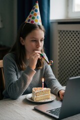 Fototapeta premium Teen girl with party hat blows horn at laptop. Birthday cake with lit candle sits on table. She celebrates alone at home, feeling sad due to isolation.