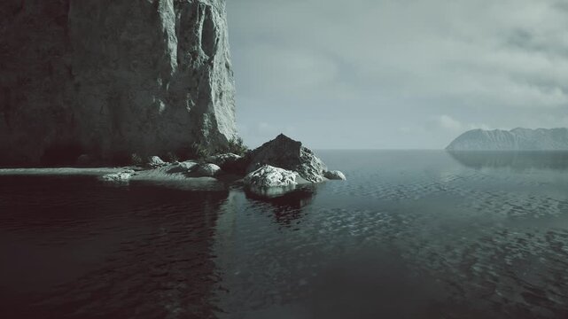 A peaceful coastal view showcases rugged cliffs and smooth waters under a cloudy sky. The shoreline features scattered rocks and subtle reflections on the waters surface.