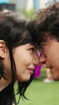 Vertical composition: Young couple touch foreheads with an intimate gaze on a picnic blanket in a city park while showing affection and flirting face to face during a romantic date.