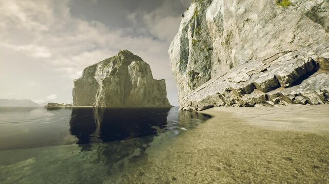 Waves gently lap against a sandy shore as sunlight casts a golden hue on majestic cliffs. The still water mirrors the rocky outcrop, creating a peaceful atmosphere at dusk.