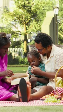 Vertical video: Mother reads an open book to a boy held by his father while they relax on a picnic blanket beside a fruit basket during shared family reading outdoors.