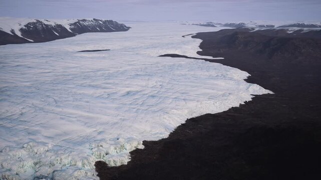 A stunning view captures the expansive glacier flowing through Greenland, surrounded by rugged mountain terrain under a clear sky. The cool, blue ice contrasts with the dark earth.