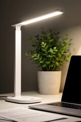 Naklejka premium Modern white desk lamp illuminates workspace with laptop, documents. Green potted plant sits on table next to computer. Minimalist home office interior with bright light for productive remote work,