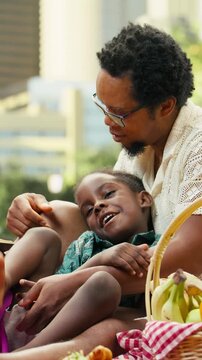 Vertical video: Mother reads a book to a boy held by his father on a picnic blanket beside a ball and fruit basket in an urban park during family bonding and storytelling.