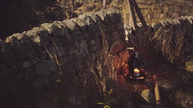 A glowing candle shines from a lantern placed on the ground near an ancient stone wall. Tall grasses surround the area, creating a serene atmosphere during twilight.