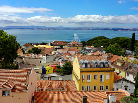 An elevated viewpoint looks over the terracotta rooftops and colorful residential facades of Lisbon toward the wide blue Tagus River and a distant ferry under a partly cloudy sky