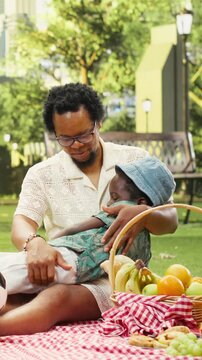 Vertical composition: Caring father holds a resting child on his lap while mother watches beside a soccer ball and picnic basket in an urban park during relaxed family time.
