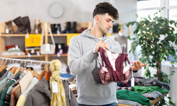 Casually dressed young guy checking lace bras in local clothing store while buying personal gift for partner