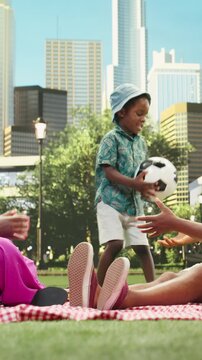 Vertical frame: Parents and boy play catch with a soccer ball beside a picnic basket, enjoying an active sunny day in an urban park with a picnic blanket, family bonding, outdoor play and quality time