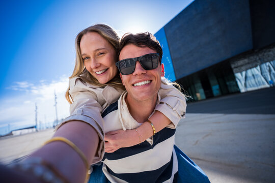 Happy young couple taking a playful selfie during a piggyback ride outdoors in a sunny modern urban setting, conveying affection, fun and youthful connection.