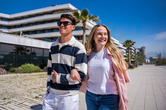 Young couple walk closely together through a sunny modern neighborhood, smiling and looking around while enjoying a relaxed urban day.