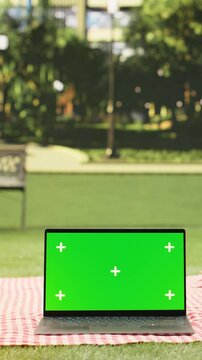 Portrait format: Green screen mockup is shown on a laptop near picnic basket with fruits and orange juice in park, screen replacement with picnic food in empty city park.