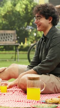 Vertical aspect: Smiling boyfriend and girlfriend say cheers and enjoy a drink on a picnic blanket, sharing affection and relaxation during an outdoor date in the city park while pouring orange juice.