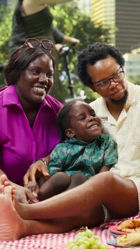 Portrait format: Smiling parents and boy laugh for a smartphone photo during a joyful family picnic in an urban park, making memories with smartphone photos during weekend fun and relaxed family time.