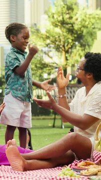 Vertical frame: Smiling boy and father exchange playful hand gestures while mother sits nearby as they play rock paper scissors and giggle on a picnic blanket in the city park.