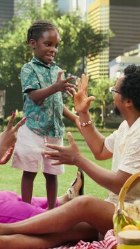 Vertical video: Smiling boy and father exchange playful hand gestures with mother nearby while playing rock paper scissors and giggling on a picnic blanket in a city park.