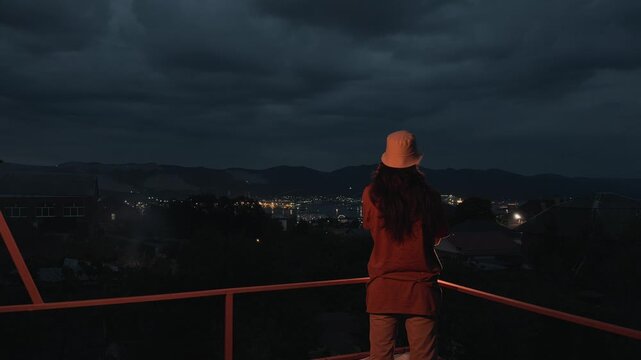 Unrecognizable anonymous long haired man in bucket hat standing on terrace with crossed arms then putting hands behind head looking at night coastal city port lights and cloudy sky rear view static