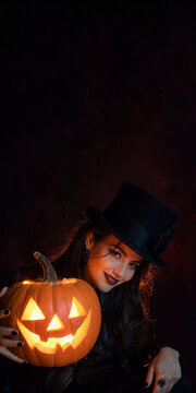 Beautiful young woman in witch top hat holding glowing jack-o-lantern pumpkin on dark background