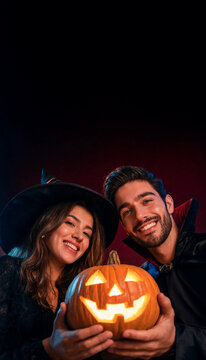 A happy smiling couple, dressed as a witch and a vampire, holding a carved glowing jack-o'-lantern together against a dark, dramatic background. Close-up portrait