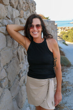 A smiling woman stands beside a stone wall with a coastal view near Mandrakia, Milos, Greece, on a sunny afternoon.