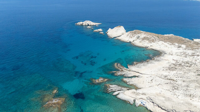 Aerial view of the rugged white coastline and turquoise sea near Mandrakia, Milos, Greece, under a bright summer sky.