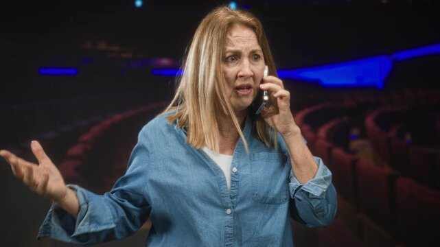 Middle aged woman in denim shirt holding smartphone to ear, gesturing with hand among red theater seats in a cinema building; frustration concern.