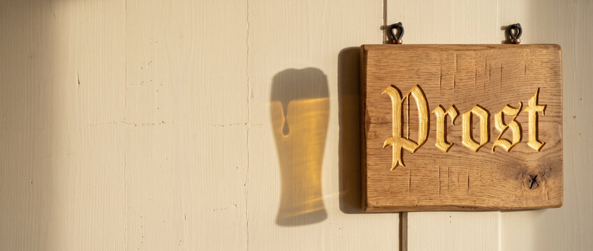 Wooden prost sign with golden gothic lettering and beer glass shadow on wall. Oktoberfest cheers and german beer festival concept. Traditional bavarian pub decor background