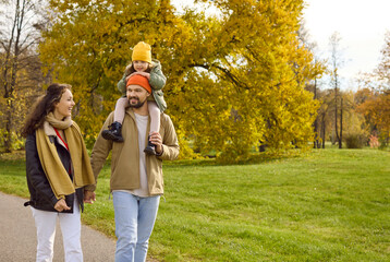 Fototapeta premium Family in autumn park, parents with child daughter. Father carries the girl on shoulders as they walk a path amid golden leaves, sharing happiness and bright smile. Warm family joy and unity.