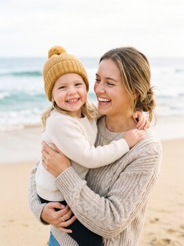 Happy mother and daughter hugging on a beach wearing cozy sweaters and a knit beanie hat.