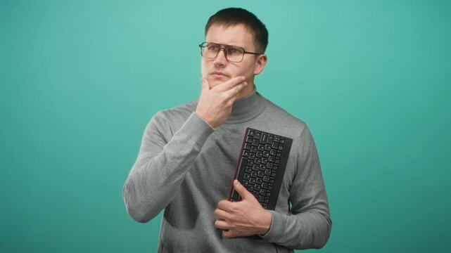 Man holding keyboard, hand on chin pensive pose wearing glasses and gray turtleneck in studio; thinking problem solving reflection.