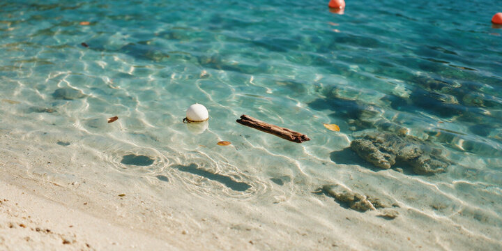 Close-up of clear shallow water with floating driftwood and white buoy. Transparent sea surface over sandy bottom with sunlight reflections