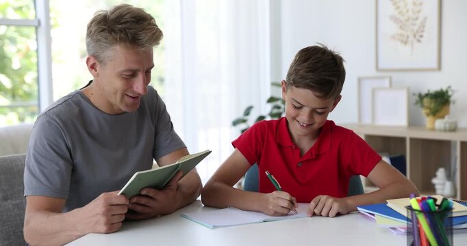 Smiling father with book helping his son with homework at white table indoors