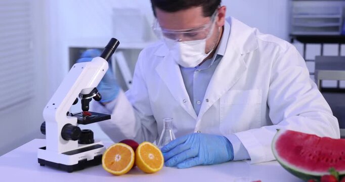 Quality control. Scientist in protective goggles, mask and gloves working at table with microscope, samples and products in laboratory. Camera moving forward