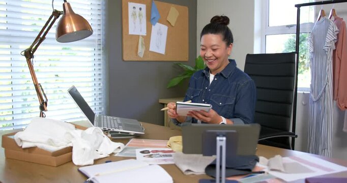 Asian woman showing notebook to laptop during call, writing while reviewing swatches in studio