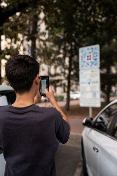 Rear view of unrecognized man scanning a parking QR code with a smartphone beside parked cars, representing mobile payment, contactless service, navigation and modern city infrastructure.