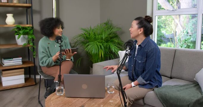 Diverse female cohosts reading tablet for podcast, speaking into mics and gesturing over laptop