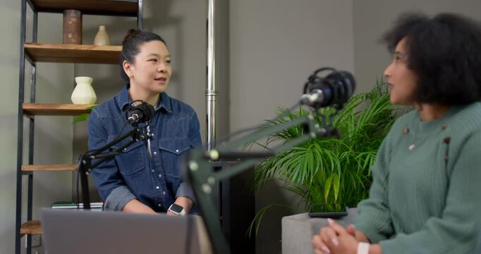 Asian, African American women recording podcast after left prompt in studio, using laptop and mics