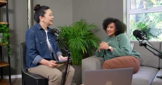 Asian host denim checking tablet, African American guest sage sweater gesturing into podcast mics
