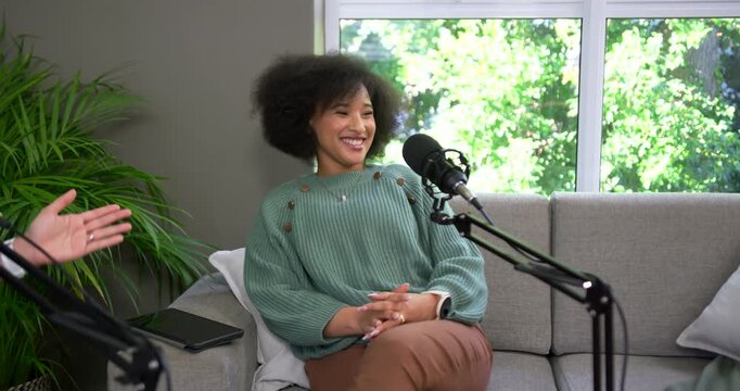 Adult woman in green top laughing, reacting to offscreen host gestures on sofa, podcast mic, tablet