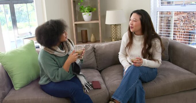Diverse female friends chatting on gray sofa, right woman leaning, reaching, taking clear glass mug