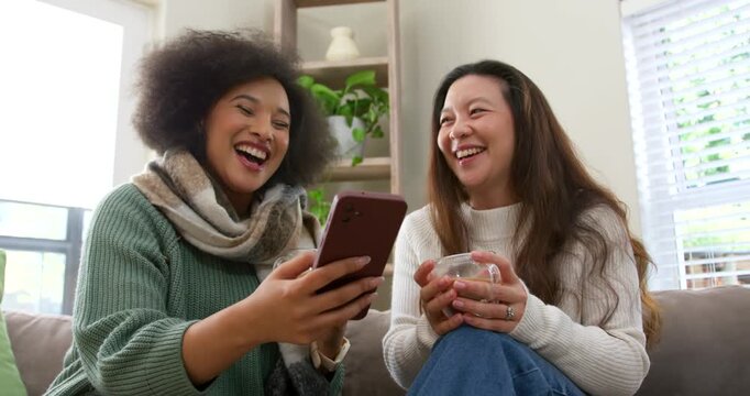 Diverse female friends in knits on couch left woman showing phone both holding glass mugs laughing