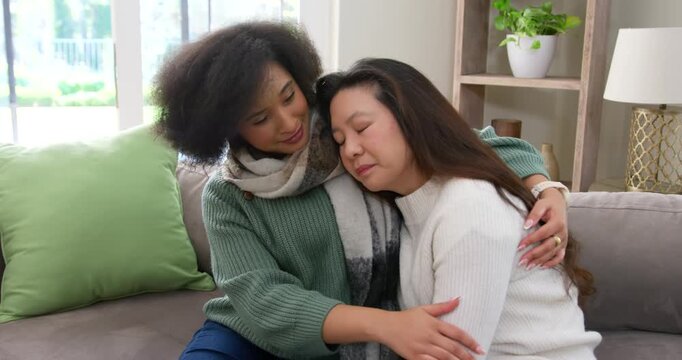 African American woman wrapping and stroking Asian woman leaning head, soothing on gray couch
