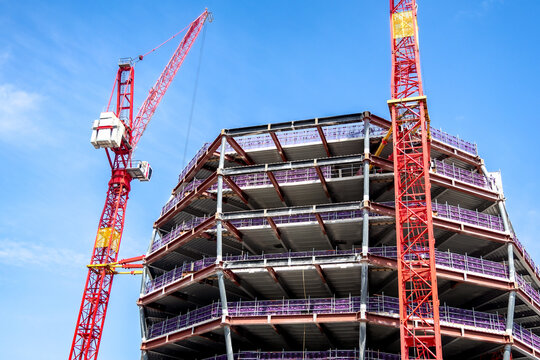 Two red tower cranes stand beside the exposed steel framework of a multistory building under construction against a bright blue sky in London, England