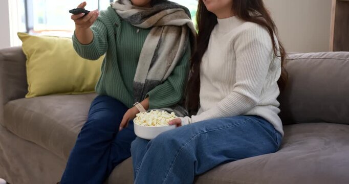 Diverse female friends sitting on sofa, left reaching remote, right holding popcorn, selecting show