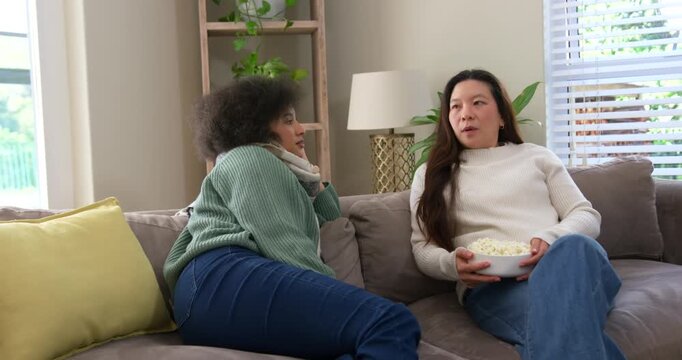 Diverse female friends talking, right woman starting story while holding popcorn bowl on gray couch