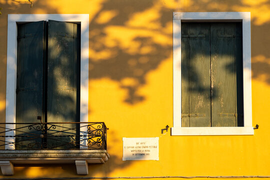 Dappled sunlight falls on yellow shutters beside a WWI memorial plaque honoring Capt. Cimbro Pettinau, Burano, Venice, Italy