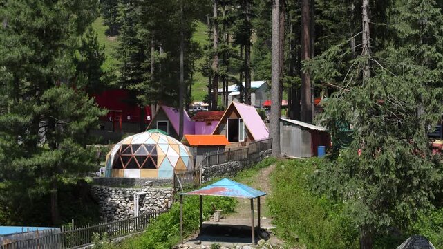 Colorful camping cabins and a geodesic dome stand among tall conifers in Kumrat Valley, Pakistan